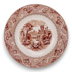 May include: A round, white ceramic plate with a brown transferware design. The center features a landscape with a castle, river, and figures in a boat. The rim has a floral and scroll pattern. A decorative piece for serving or display.