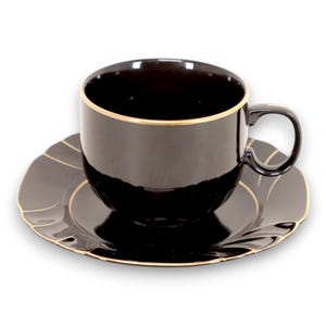Puede incluir: Juego de taza y platillo negro con detalles dorados. La taza tiene un asa redondeada y se apoya en un platillo a juego con borde festoneado. El ribete dorado acentúa el borde de la taza y el borde del platillo.