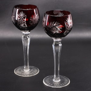 Puede incluir: Dos copas de vino con cuencos de color rojo intenso y patrones geométricos y de estrella tallados en el cristal. Cada copa tiene un tallo transparente facetado y una base circular con un diseño radial, mostrando un contraste entre el vidrio rojo oscuro y los elementos de cristal.
