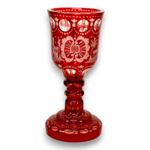 May include: A ruby red goblet with detailed white etched patterns. The glass has a floral and architectural design around the bowl and base. The stem is ribbed, and the foot is round, creating an elegant, vintage aesthetic.