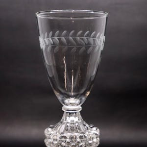 May include: A clear glass goblet with a decorative leaf pattern etched around the top. The stem features a textured, bubble-like design. The glass is designed to hold liquids.
