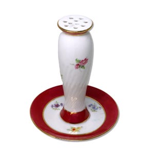 Könnte beinhalten: Eine weiße Porzellan-Blumenhalterung mit rotem und goldfarbenem Rand. Die Blumenhalterung hat eine zylindrische Form mit Löchern an der Oberseite. Der Sockel ist eine Platte mit rotem Rand und floralen Akzenten. Die Blumenhalterung ist mit rosa und lila Blumen verziert.
