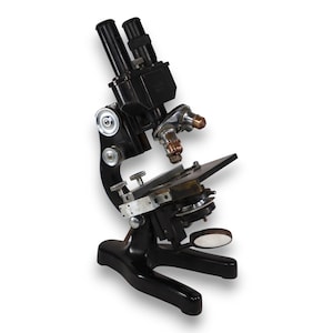 May include: A vintage black microscope with dual eyepieces, a stage, and adjustable knobs. The microscope has a classic design with a sturdy base and is set against a white background. The lenses and metal components show signs of age.