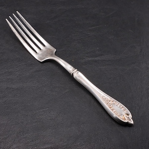 May include: A silver fork with four tines and a decorative handle. The handle features an ornate floral design near the base and a smooth, curved shape. The fork is set against a dark background.