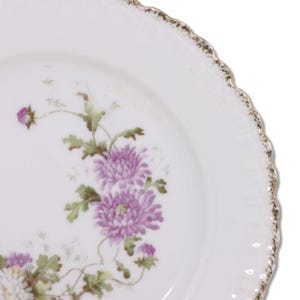 Puede incluir: Un plato de porcelana blanca con un borde festoneado y un borde dorado. El plato presenta un diseño floral con flores rosas y verdes.