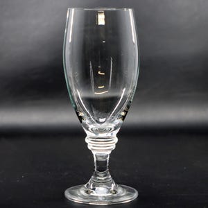 May include: A clear, stemmed glass with a tulip-shaped bowl and a round base. The stem features a decorative, ridged design. The glass is designed for beverages and is set against a dark background.
