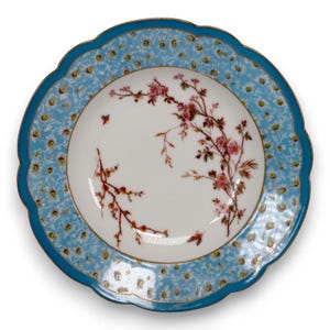 May include: A round porcelain plate with a scalloped edge featuring a white center decorated with delicate pink floral branches and small insects. The wide border is light blue with a repeating pattern of white daisies and a gold rim.