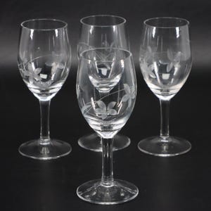 May include: A set of four clear glass wine glasses with a slender stem and a circular base. Each glass features a delicate, etched floral pattern consisting of five-petaled flowers and thin, curved stems wrapping around the bowl of the glass.