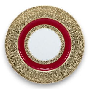 May include: A round, decorative plate with a white center, a vibrant red band, and an ornate gold border. The gold border features a repeating pattern of stylized leaves and a beaded edge. The plate is likely made of porcelain.