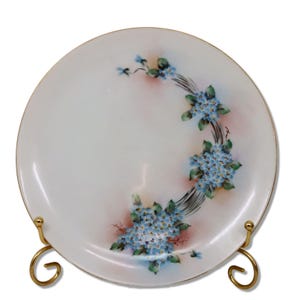 Könnte beinhalten: Ein weißer Keramikteller mit Goldrand, verziert mit einem Blumendesign aus blauen Vergissmeinnicht und grünen Blättern. Die Blumen sind in einem gebogenen Muster angeordnet. Der Teller wird auf einem goldfarbenen Metallständer präsentiert.