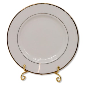 May include: A white plate with a gold rim and a thin black line inside the rim. The plate is displayed on a gold-colored metal stand with a decorative, scrolled design. The plate is round.
