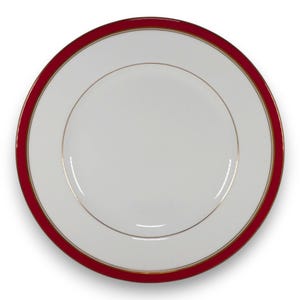 May include: A round white porcelain dinner plate featuring a wide, solid deep red rim. The plate is accented with a thin, circular gold band separating the white center from the red border, and a second gold band along the outer edge of the rim.