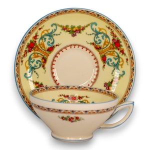May include: A vintage teacup and saucer set. The cup and saucer are cream-colored with a yellow center and blue trim. The set features a decorative design of fruit, flowers, and ornate scrollwork in red, blue, and gold.