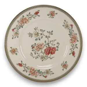 May include: A round, off-white ceramic plate with a floral design. The plate features a border with a repeating pattern and a gold rim. The floral design includes pink and peach-colored flowers with green leaves. The plate is a decorative item.