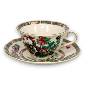 May include: A cream-colored porcelain teacup and saucer set featuring a vibrant floral pattern with pink, red, and green blossoms on brown branches. The rim of both pieces is decorated with a dark brown geometric border pattern.