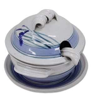 May include: A ceramic lidded bowl with a matching saucer and a small ladle. The off-white glazed pottery features hand-painted blue and teal brushstroke patterns of grass and flowers. The lid has a decorative curled handle and a side notch.