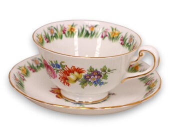 May include: A white teacup and saucer set with a floral design. The cup and saucer have a gold trim. The floral design includes red, yellow, purple, and blue flowers. The rim of the cup has a green and yellow grass design.