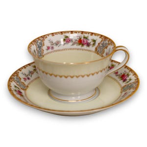 Könnte beinhalten: Cremefarbene Teetasse und Untertasse mit floralen und goldenen Verzierungen. Die Tasse hat eine runde Form mit Henkel, die Untertasse ist kreisförmig. Das Set zeigt ein zartes Blumenmuster mit rosa und roten Rosen.