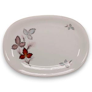 May include: An oval white ceramic serving platter featuring a botanical illustration of leaves in shades of red, brown, and muted green on the left side, with smaller leaf accents positioned on the upper right and lower right edges.