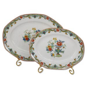 May include: Two white ceramic serving platters with floral designs and decorative borders. The platters are oval and octagonal, featuring colourful flowers and a green, blue, and red border. They are displayed on gold-coloured stands.