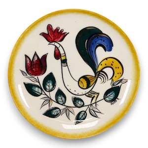 May include: A decorative ceramic plate with a rooster design. The plate features a white base with a yellow rim. The rooster is depicted with red, blue, yellow, and green accents. A red flower and green leaves are also present.