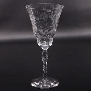 May include: A clear crystal wine glass featuring an intricate etched floral pattern on the bowl. The stem has a faceted, twisted design that leads down to a circular base with a scalloped edge. The glass is transparent with reflective surfaces.