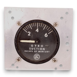 May include: A vintage gyro suction gauge with a black face and white markings, mounted on a silver metal plate. The gauge reads "2", "4", and "6" with the text "INCHES OF MERCURY" below. Four screws secure the gauge to the plate.