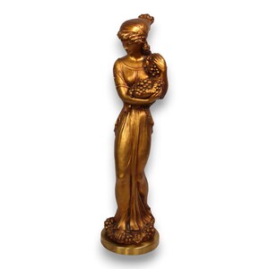 May include: A gold-colored statue of a woman holding grapes. The figure is wearing a draped gown and a head covering. The statue is standing on a round base.