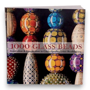 May include: The book cover for "1000 Glass Beads" displays a collection of colourful glass beads against a dark backdrop. The title is accompanied by the subtitle "Innovation & Imagination in Contemporary Glass Beadmaking." The beads showcase diverse shapes and patterns, including orange, blue, and yellow.