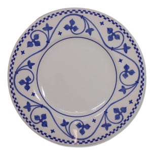 May include: White ceramic plate with a blue floral design. The plate features a central white area, a blue border with a checkered pattern, and a decorative blue floral and scroll design around the edge. The plate is round.
