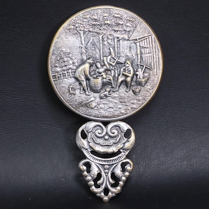 May include: A silver-toned, round decorative piece with a detailed relief scene of people in a village setting. Below, an ornate, heart-shaped embellishment with floral and scroll designs. The piece is set against a black background.