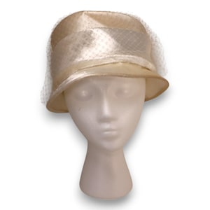May include: A vintage cream-colored hat with a rounded crown and a small brim. The hat features a satin band and a delicate net veil. The hat is displayed on a white mannequin head, showcasing its shape and design.