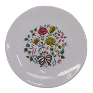 Può includere: Piatto in ceramica bianca con un motivo a bouquet floreale. Il bouquet presenta fiori rossi, gialli e verdi, con un fiocco marrone alla base. Il piatto ha un design semplice ed elegante.