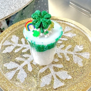 May include: A St. Patrick's Day themed dessert in a clear cup, featuring layers of white and green. It's topped with a glittery green shamrock, a rainbow, and white sprinkles. The dessert sits on a gold glittery tray with a snowflake design.
