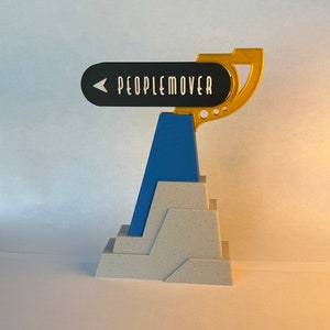 May include: A white and gray model of a building with a blue and yellow sign that reads "PEOPLEMOVER" on top.