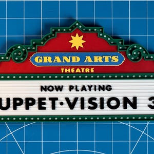 May include: A colorful theater marquee sign with the text "GRAND ARTS THEATRE" and "NOW PLAYING MUPPET-VISION 3D". The sign has a red and green border with a yellow star and a white marquee.