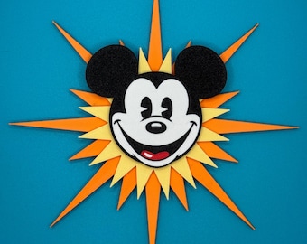 Puede incluir: Una decoración de sol con el tema de Mickey Mouse. La imagen central presenta la cara de Mickey con orejas negras, una cara blanca y una boca roja. El sol tiene rayos naranjas y amarillos sobre un fondo azul.