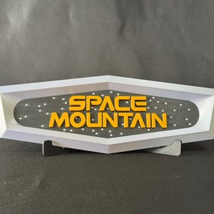 以下が含まれることがあります： オレンジ色のブロック体で「SPACE MOUNTAIN」と書かれた3Dプリントのサイン。サインは白で、内側は黒、小さな白い点があります。黒いスタンドで支えられています。