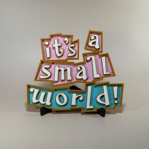 以下が含まれることがあります： 白い文字で「it's a small world!」というフレーズが入ったカラフルなサイン。 ピンク、ティール、ゴールドの配色で、黒いスタンドで支えられています。