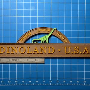 Op de afbeelding: Een bruin bord met de tekst "DINOLAND U.S.A." in gouden letters. Boven het bord staat een groen dinosaurus silhouet en een bruine boog met de woorden "EXPLORATION", "EXCAVATION" en "EXULTATION".