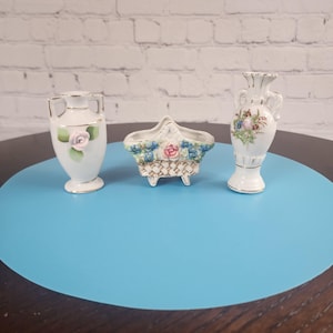 May include: Three small, decorative porcelain vases. One is a white vase with a pink rose and gold trim. Another is a basket-shaped vase with blue and pink floral designs. The third is a white vase with floral designs and gold trim.
