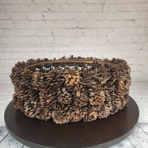 May include: A round, decorative basket crafted from numerous brown pine cones. The pine cones are densely packed, creating a textured surface. The basket sits on a dark wooden surface, with a white brick wall in the background.