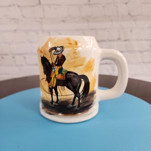 May include: White ceramic mug with a brown and black illustration of a cowboy riding a horse. The cowboy is wearing a brown hat and yellow pants. The mug has a handle.