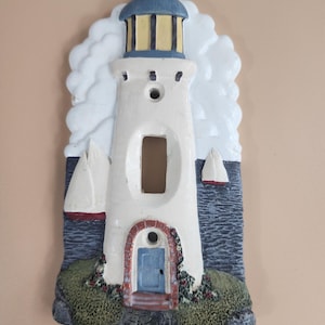 May include: A ceramic light switch cover featuring a white lighthouse with a blue roof and a yellow window. The lighthouse is surrounded by blue water, white clouds, and green grass. There are two sailboats in the water.