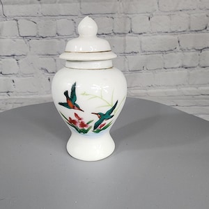 May include: White porcelain jar with a lid, featuring a floral design and two green and blue birds in flight. The jar has a gold trim around the lid and base.