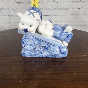 May include: A blue and white porcelain figurine of two pigs laying on a log. The pigs are wearing blue and gold clothing. The figurine is on a wooden surface.