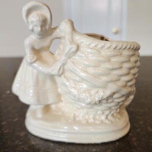 May include: White ceramic planter shaped like a girl carrying a basket. The planter is a vintage style and has a delicate design.
