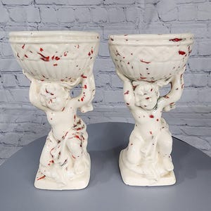 May include: Two white ceramic planter stands with red speckles. Each stand features a cherub figure holding up a bowl-shaped planter.
