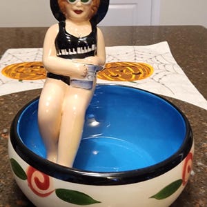 May include: A ceramic bowl with a blue interior and a white exterior. The bowl is decorated with red and green floral designs. A ceramic figurine of a woman wearing a black swimsuit and sunglasses is sitting on the rim of the bowl.