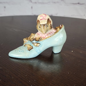 May include: A light blue ceramic shoe figurine with a mouse and its babies inside. The mouse wears a pink and white checkered dress and a matching bonnet. The shoe has a high heel and decorative cutouts.